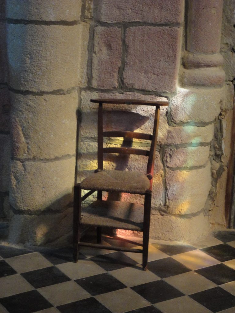 A chair stands against a stone wall. Light from a stained glass windows falls across it.